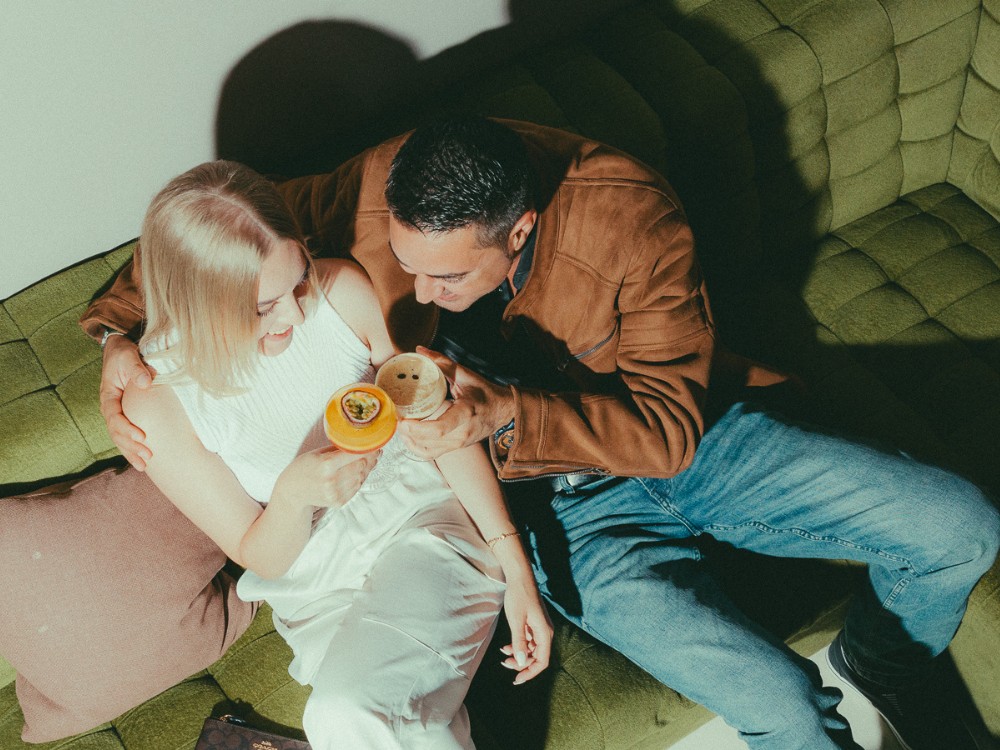 A couple sits closely on a green sofa, smiling and holding drinks. She wears a white outfit; he wears a brown jacket and jeans. The mood is joyful and relaxed.