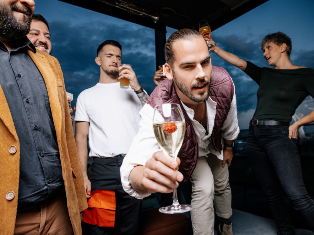 A lively group gathers at a rooftop party at dusk. A man in the foreground holds a wine glass, leaning in with a friendly gesture. Others in casual attire cheer with raised glasses, creating a festive atmosphere. The sky is a deep blue, adding to the vibrant mood.
