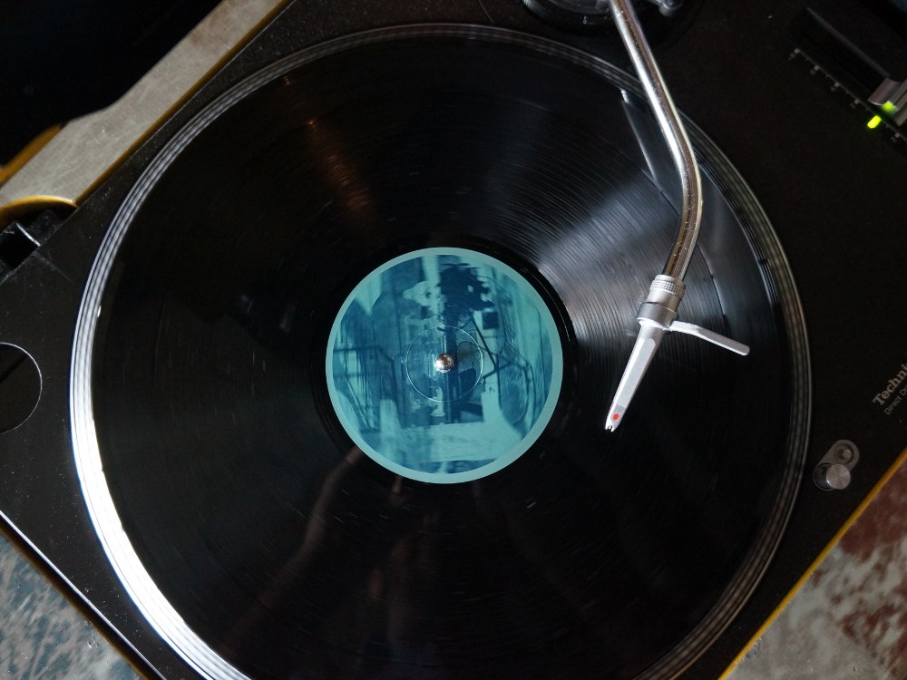 Close-up of a spinning vinyl record on a turntable, featuring a blue label and a silver tonearm. The image evokes a nostalgic, vintage atmosphere.