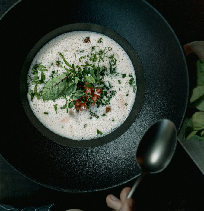 A dark bowl of creamy soup topped with fresh herbs, diced tomatoes, and microgreens, with a spoon poised nearby.