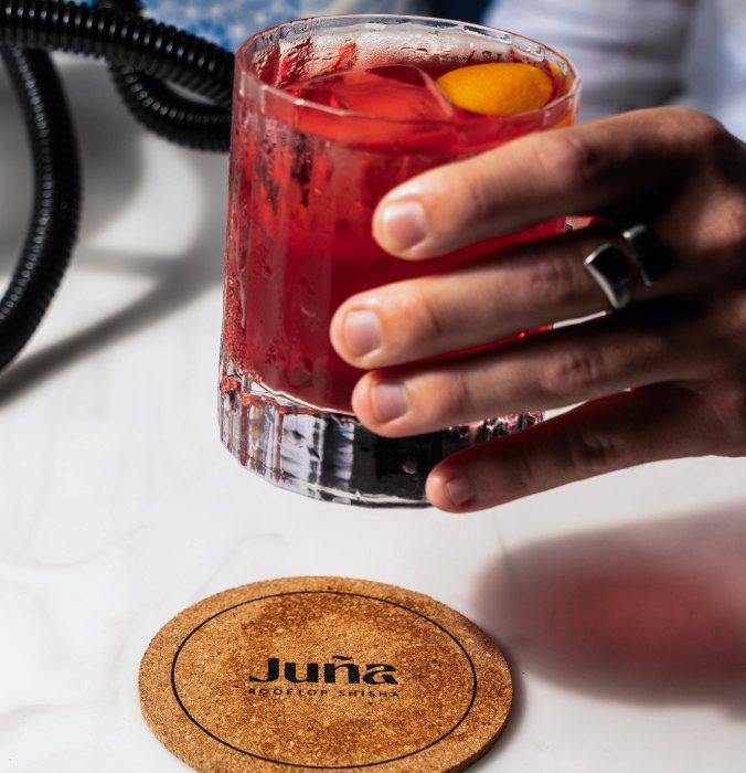 A hand holds a glass of vibrant red cocktail with ice, resting on a cork coaster that reads "Junā" against a sleek tabletop backdrop.