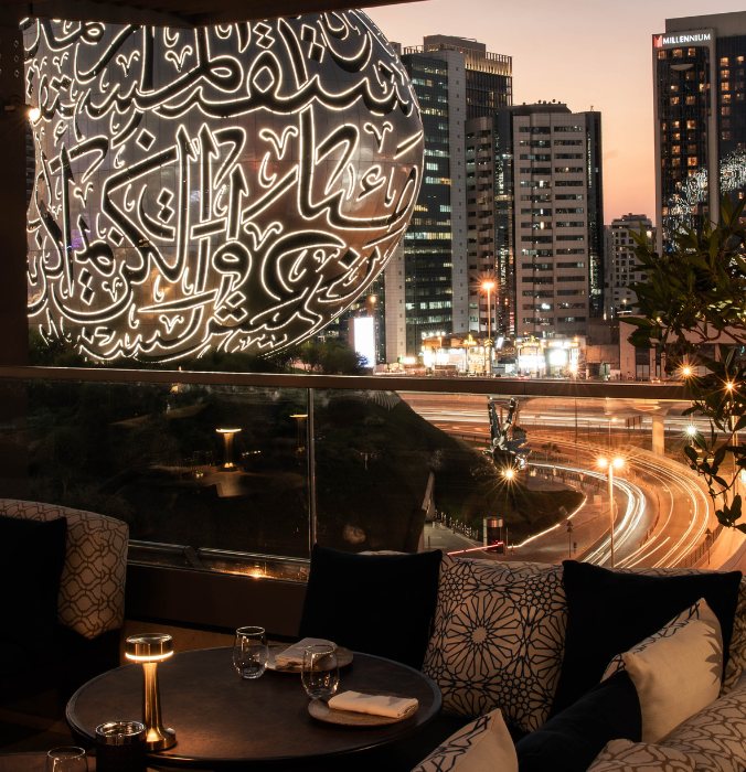 Evening cityscape of tall buildings and a round structure with illuminated Arabic calligraphy. Foreground features a cozy dining area with soft lighting.