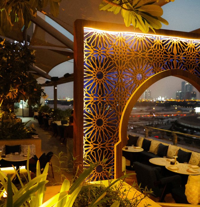 Elegant rooftop terrace with intricate lit arch design, lush green plants, and city skyline in the background at dusk. Cozy and inviting atmosphere.