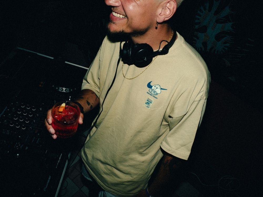 A smiling DJ wearing headphones around his neck holds a red drink while standing at a mixing console in a dimly lit setting.