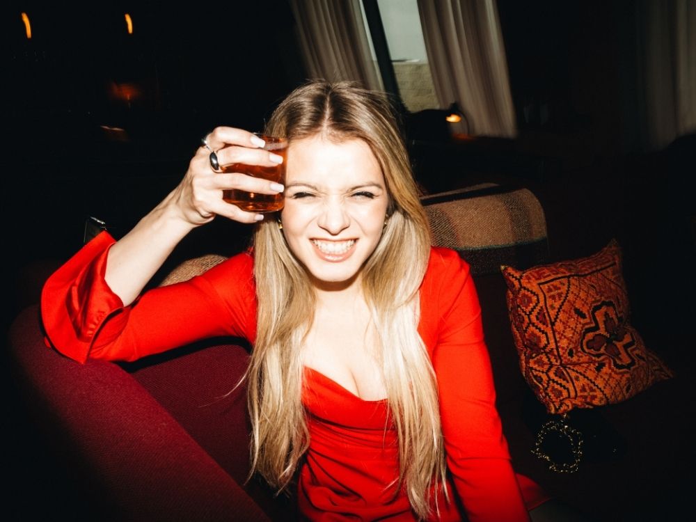A woman in a red dress laughs and holds a drink playfully to her forehead while sitting on a sofa in a dimly lit lounge.
