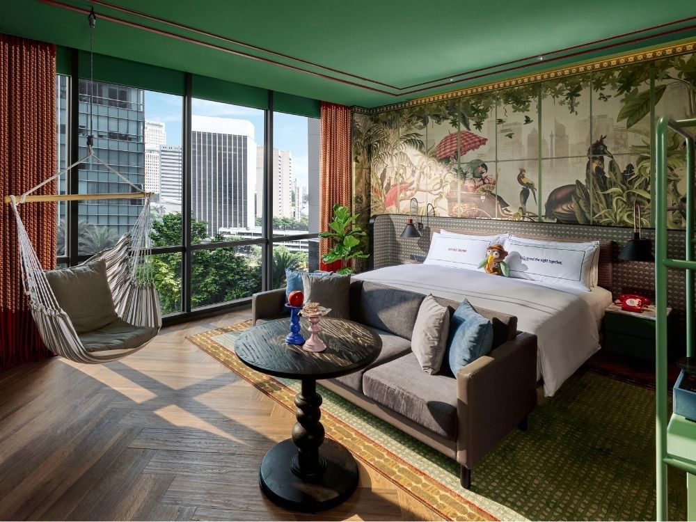 Stylish hotel room with a jungle-themed mural, large windows, and city view. Features a comfy swing chair, elegant bed, and a cosy seating area.