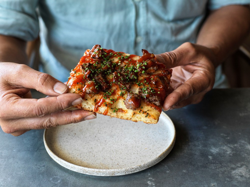 A person holds a slice of thick-crust pizza over a small, empty plate. The pizza is topped with sausage, cheese, and herbs, conveying a savory, appetizing tone.