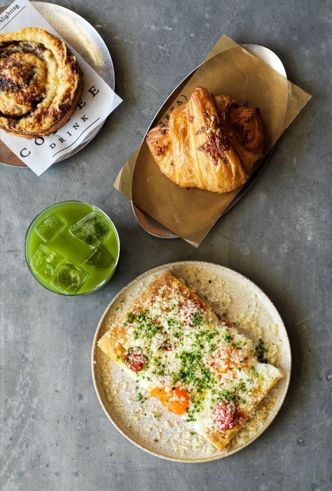 A savory pastry topped with cheese, chives, and cherry tomatoes on a beige plate, next to a green iced drink and two pastries on metal trays.