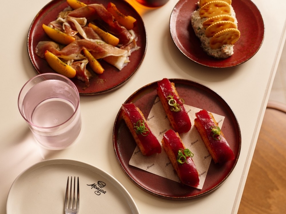 Plates of gourmet appetizers on a table. Top-left: ham and peach slices, top-right: crackers on spread, bottom: red-topped bites with garnish, next to a pink glass.