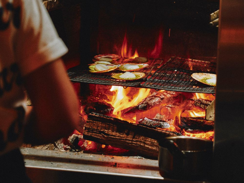 Grilled seafood sizzling over an open flame, with flames licking the wood below, as a cook monitors the cooking process.