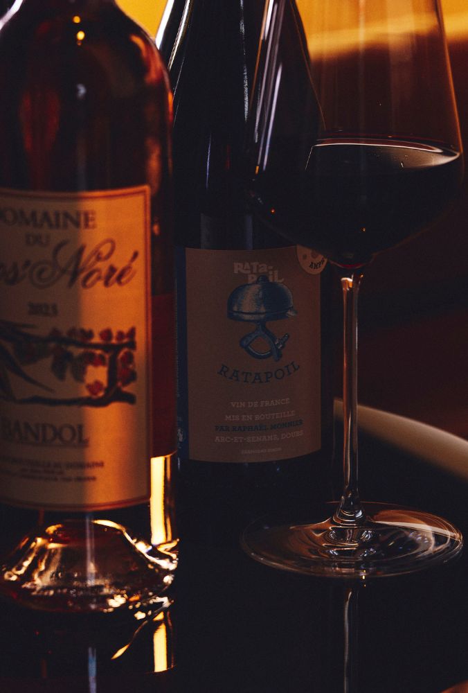 Two wine bottles with ornate labels, "Domaine du Gros' Noré" and "Ratapoil," stand beside a full wine glass. Warm, moody lighting enhances elegance.