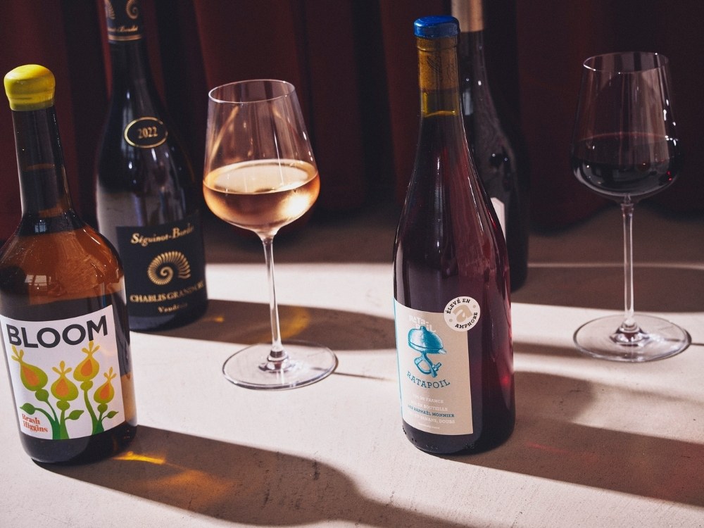 Five wine bottles and two glasses on a table. The glasses contain red and rosé wine. Sunlight creates warm shadows, suggesting an elegant setting.