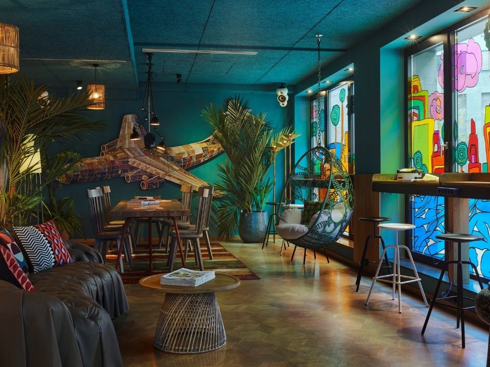 A colorful, modern lounge with an airplane mural, greenery, a wooden dining table, and vibrant stained glass windows.