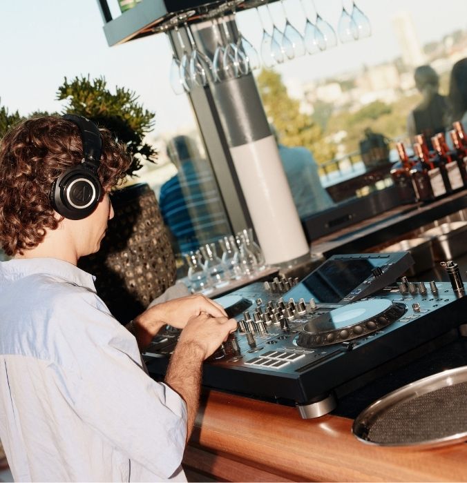 A DJ wearing headphones mixes music at a stylish outdoor bar, surrounded by glassware and a city view in the background.