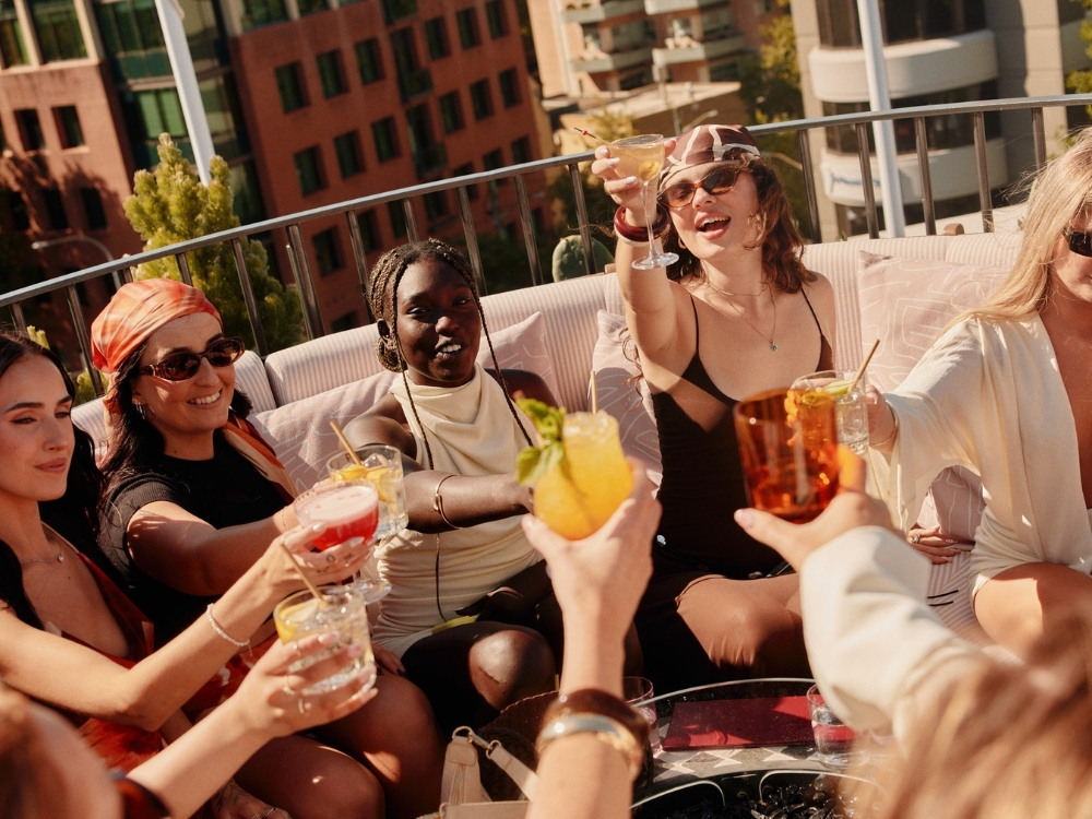 A lively rooftop gathering with friends raising colorful cocktails, set against a backdrop of modern buildings and greenery.
