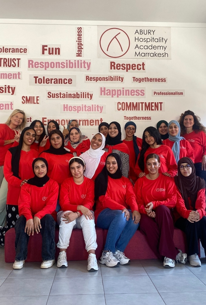 A diverse group of women in red shirts smiling confidently in front of a wall with positive words like "happiness," "commitment," and "empowerment."