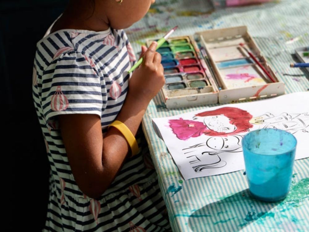 A child in a striped dress paints a colorful cartoon character using watercolors at a table. Bright and creative atmosphere with vibrant colors.