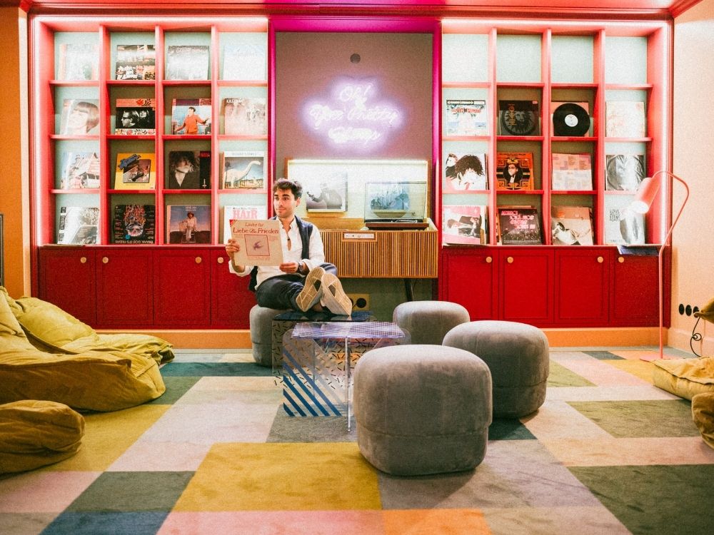 Man sitting in the colourful Vinyl Lounge at 25hours Hotel Indre By, holding a vinyl record in front of red shelving filled with albums. Soft seating, beanbags and warm lighting create a relaxed, retro-inspired atmosphere.