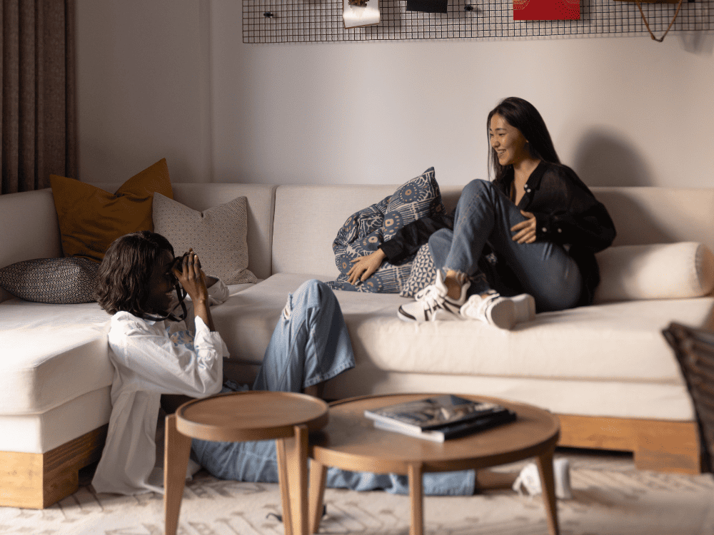 Two individuals relax on a couch in a cozy living room, one taking a photo while the other lounges with a patterned blanket.