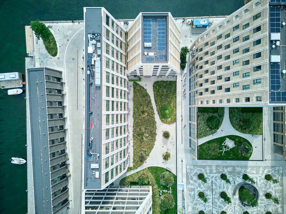 Aerial view of modern buildings surrounding a landscaped courtyard, featuring greenery, pathways, and a nearby waterway with boats.