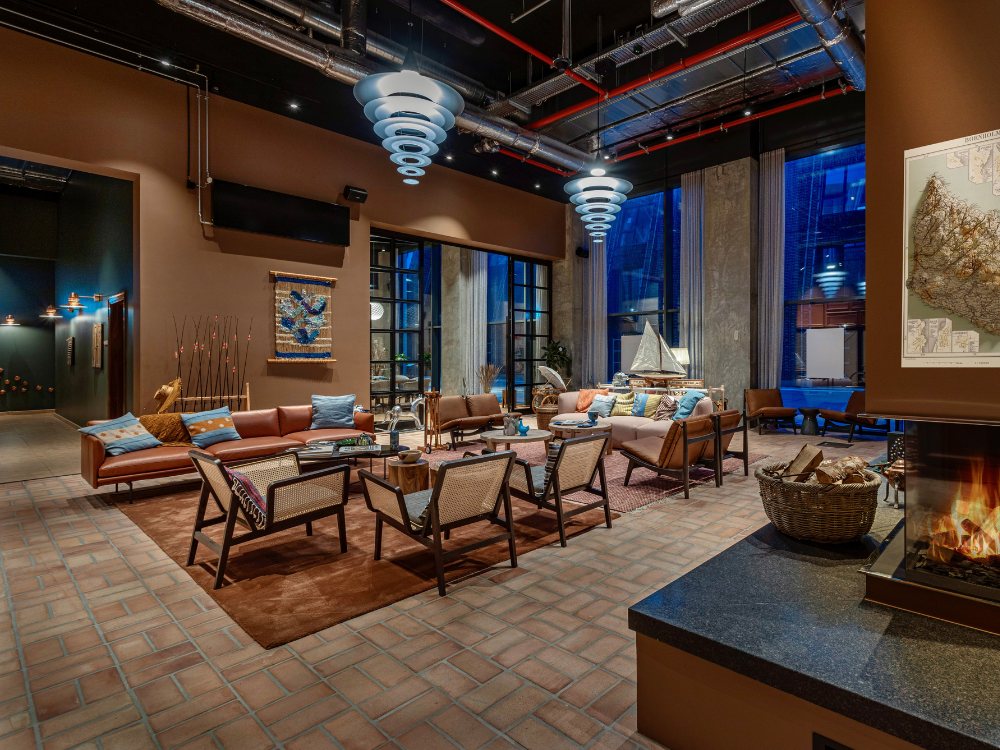 Cozy lounge featuring modern furniture, warm lighting, and a fireplace, with large windows showcasing a nighttime city view.