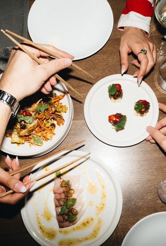 A table with various Asian dishes. Hands with chopsticks reach for plates of sushi, sliced fish, and crispy vegetables, amidst elegant dining.