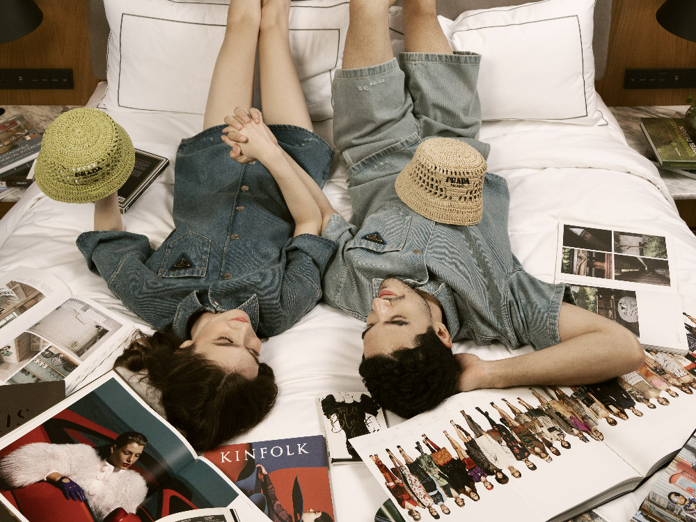 A couple in denim outfits lies on a bed, holding hands. Open fashion magazines surround them, evoking a relaxed, stylish atmosphere.