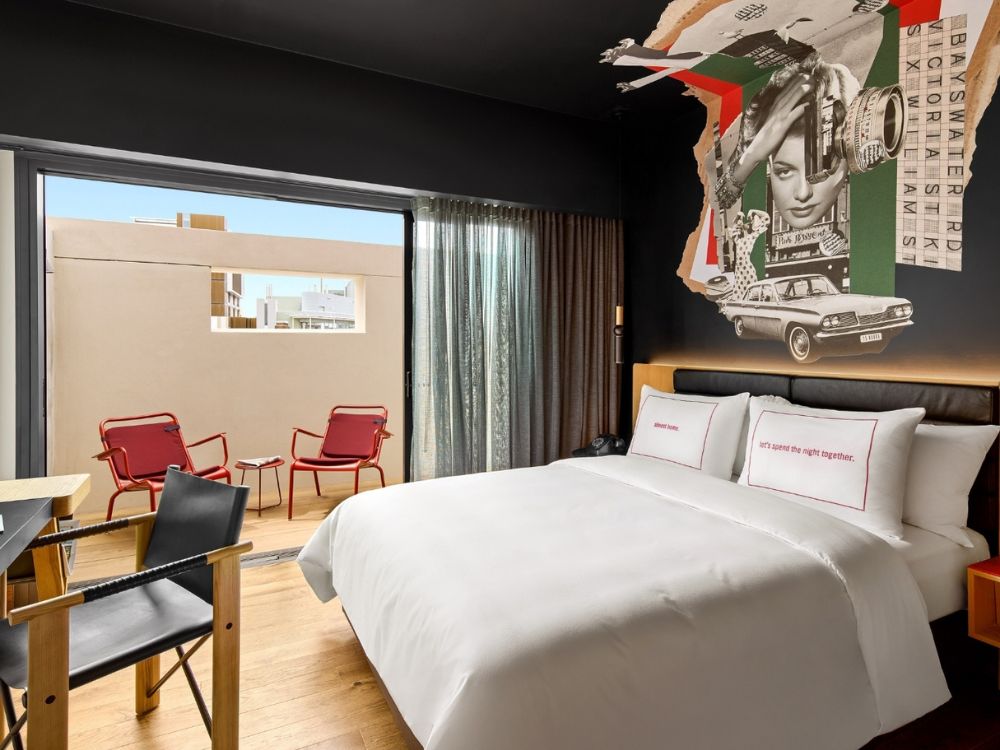 Modern hotel room with a large bed, pillows embroidered with text, and retro-style wall art. A balcony with red chairs and a small table is visible through glass doors.