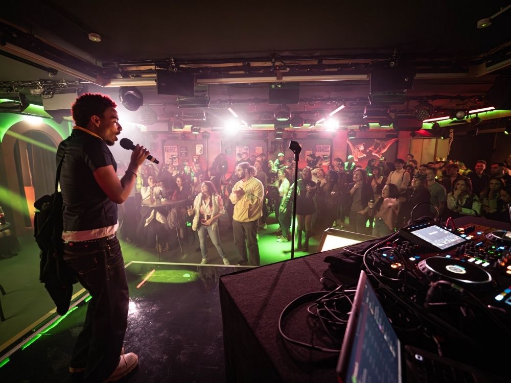 A lively crowd enjoys a performance in a dimly lit venue, with colorful lights and a DJ setup visible in the foreground.