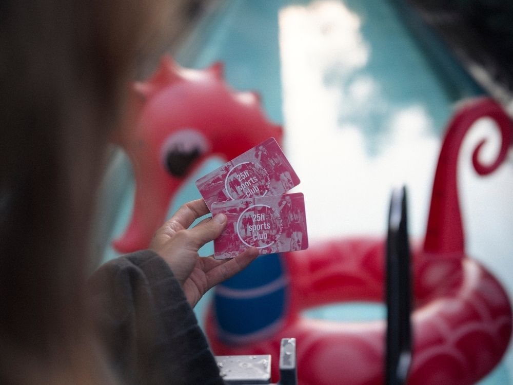 A hand holds two membership cards labeled "25h Sports Club" in front of a swimming pool. A pink, inflatable sea creature floats in the water.