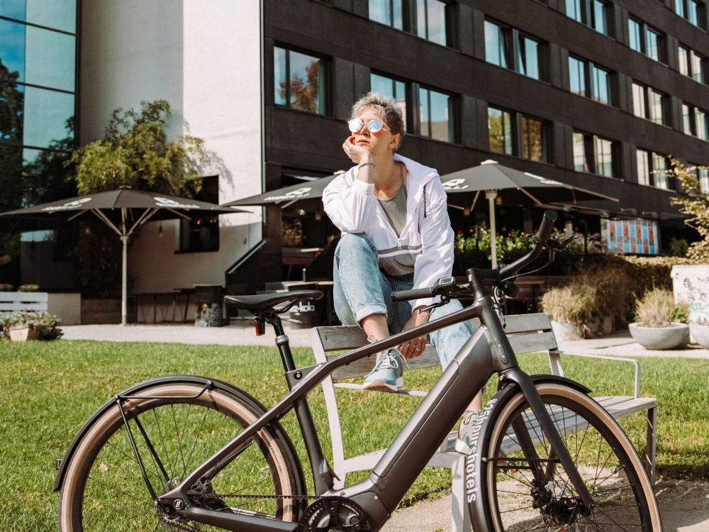 Person in casual clothes sitting on a bench beside a modern electric bike in an urban park area with building and umbrellas in the background.