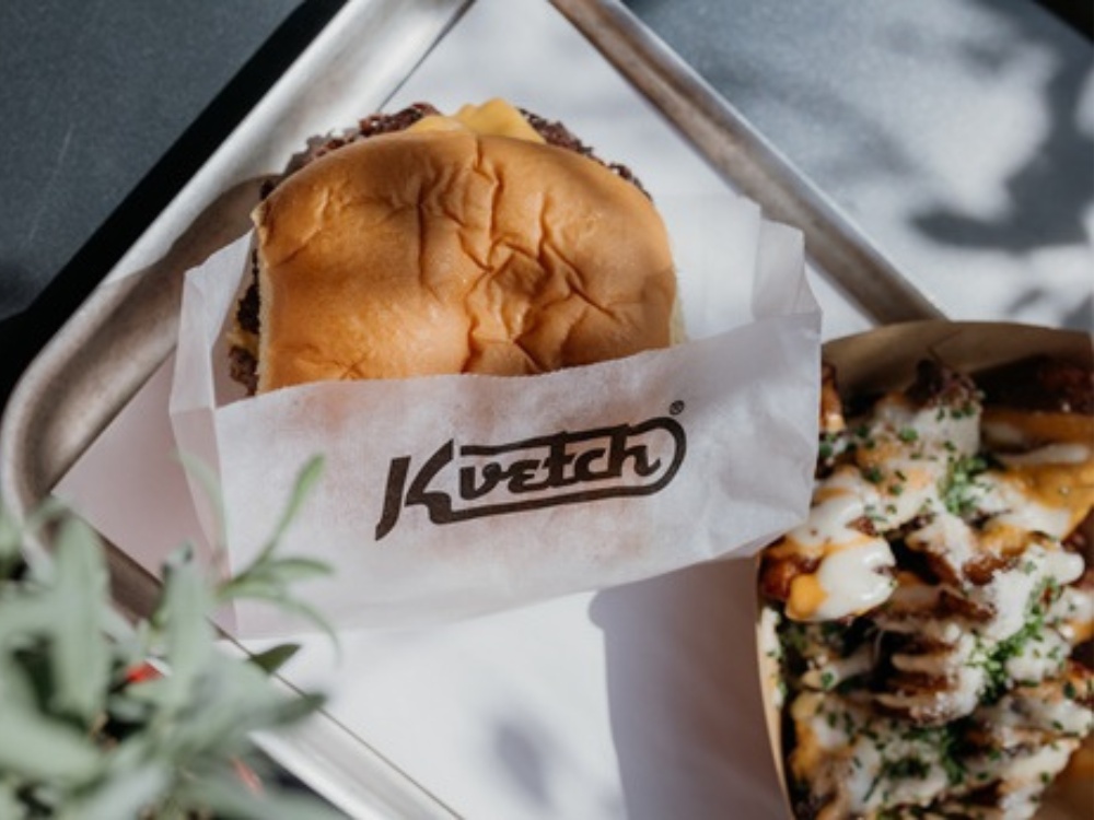 Burger wrapped in branded "Kvetch" paper on a tray beside loaded fries topped with sauce and herbs, set in bright sunlight for a fresh, appetizing look.