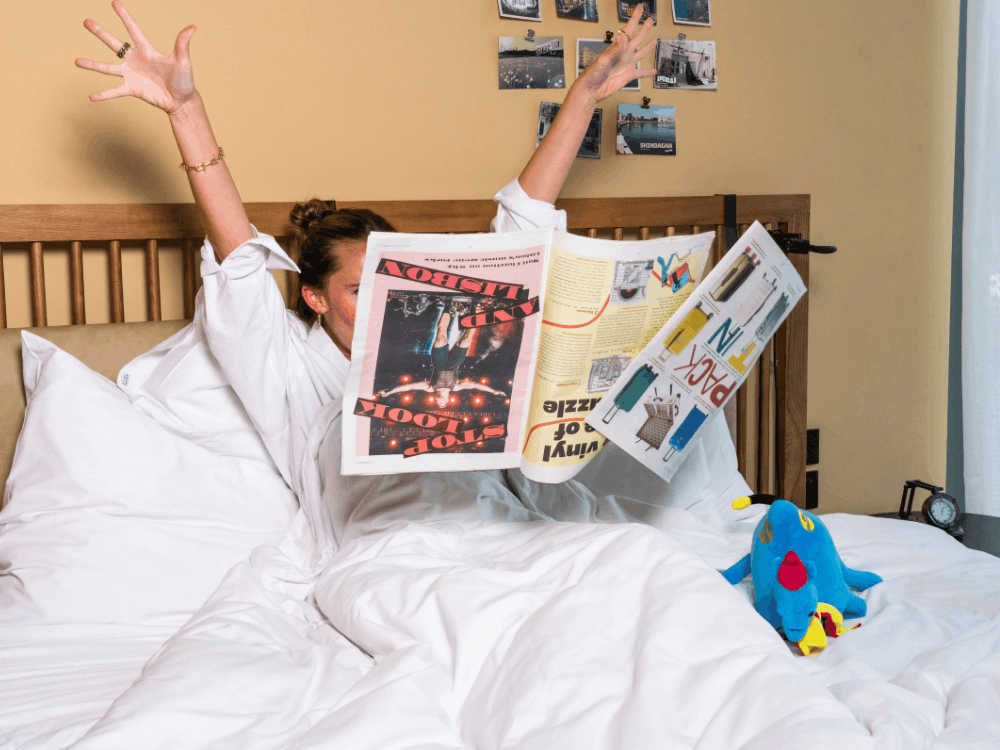 Person in bed tossing a newspaper in the air with arms raised, surrounded by white bedding and a blue plush toy on the bed.