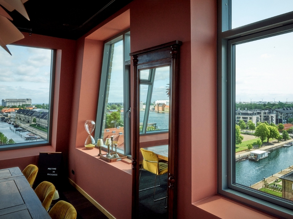 Modern room with salmon-colored walls featuring large windows showing a vibrant cityscape and river. A floor mirror reflects elegant decor. Peaceful ambiance.