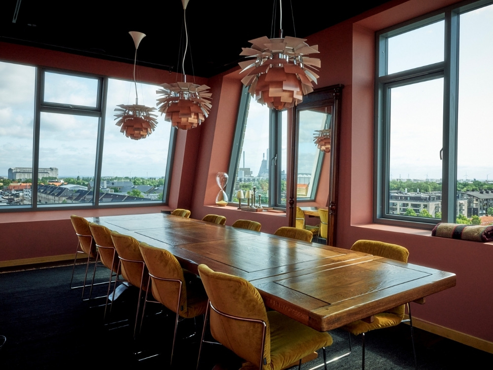 A modern meeting room with a wooden table, surrounded by yellow chairs, vibrant pendant lights, and large windows showcasing an urban skyline.