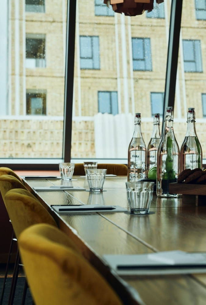 A meeting room with a wooden table, surrounded by yellow chairs. Bottles and glasses are neatly arranged. Large windows display a city building view.