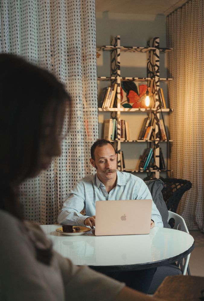 Man working on a laptop at a round table with a plate of brownies, bookshelf and curtains in a cozy, dimly lit room.