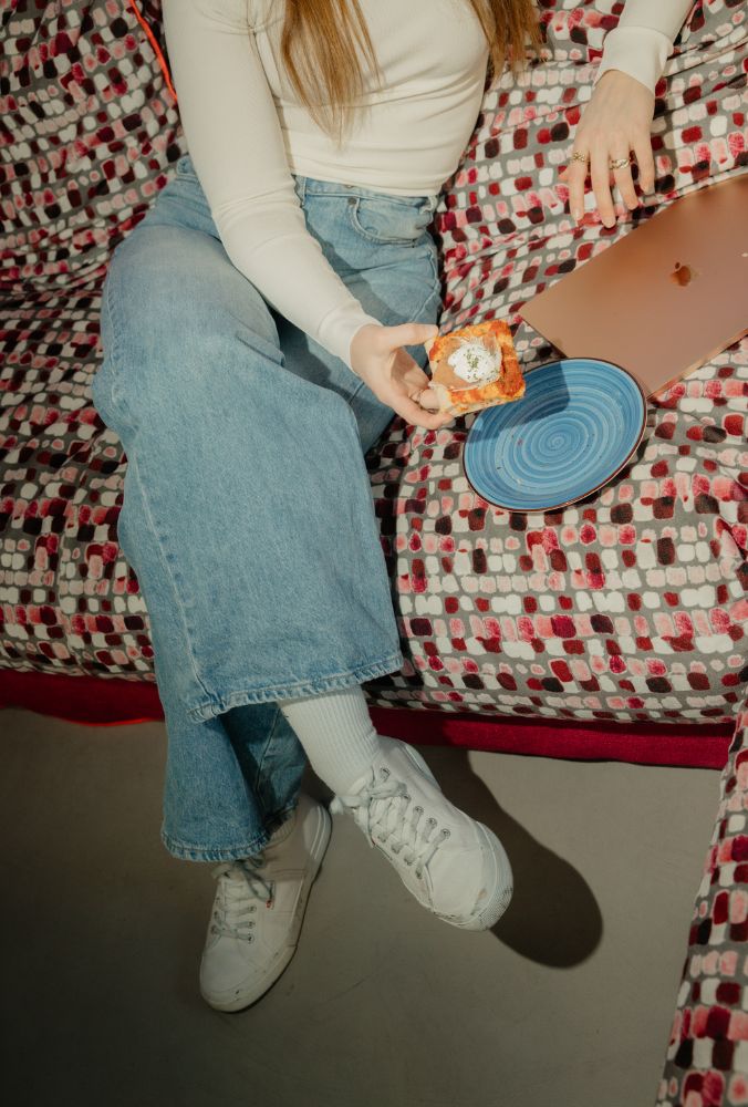 Person in jeans and white sneakers holding a slice of pizza, sitting on a patterned couch with a blue plate and laptop nearby.