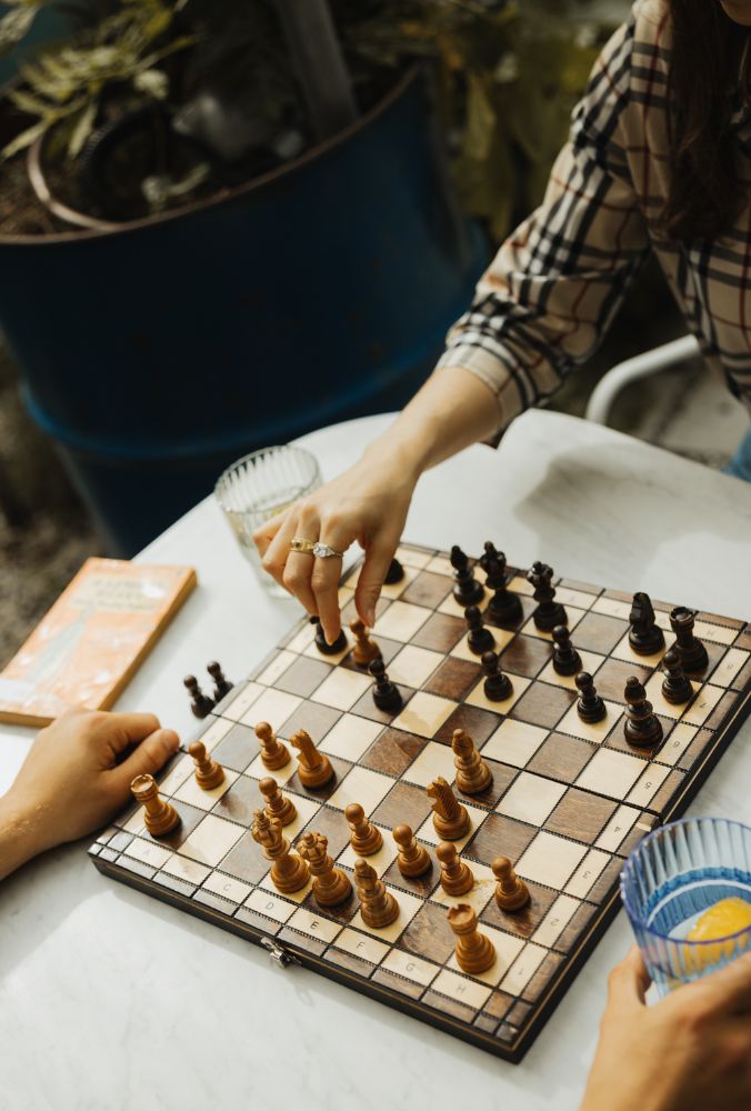 Two people playing chess at a table, one hand moving a piece, drinks and a book nearby.