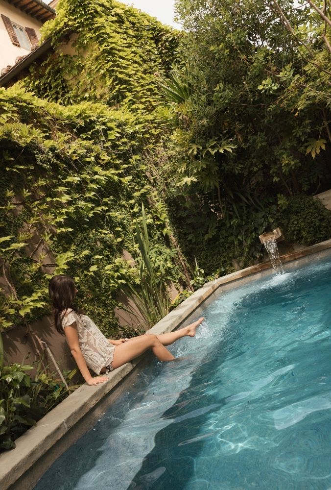 Woman sitting on the edge of a pool, dipping her feet in water, surrounded by lush green plants and ivy-covered walls.