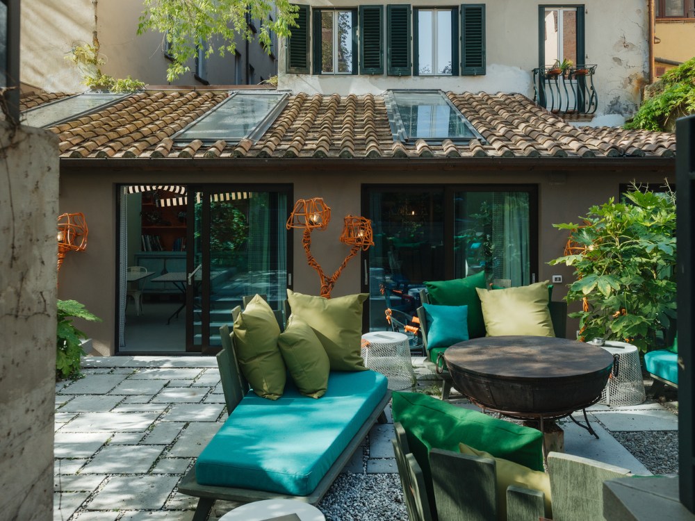 Charming patio with green and blue cushions on lounge chairs, surrounded by lush plants. Cozy, rustic vibe with clay roof tiles and large glass doors.