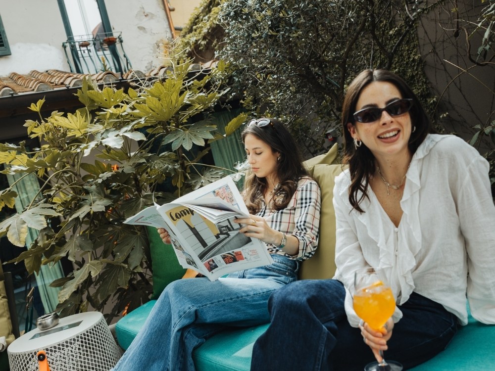 Two women relaxing on a patio sofa surrounded by lush plants, one reading a magazine and the other holding an orange cocktail.