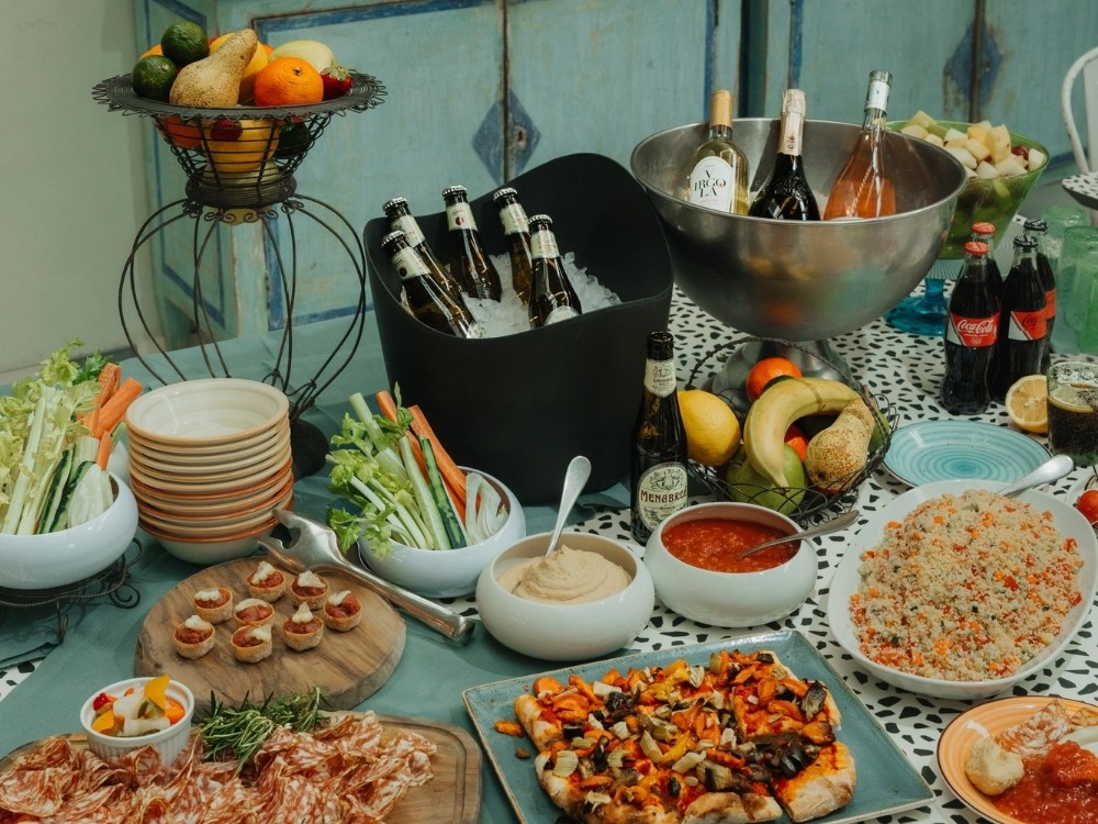 Buffet table with assorted fruits, sliced meats, pizza, dips, vegetable sticks, quinoa salad, beer bottles, wine, and soda on a patterned tablecloth.