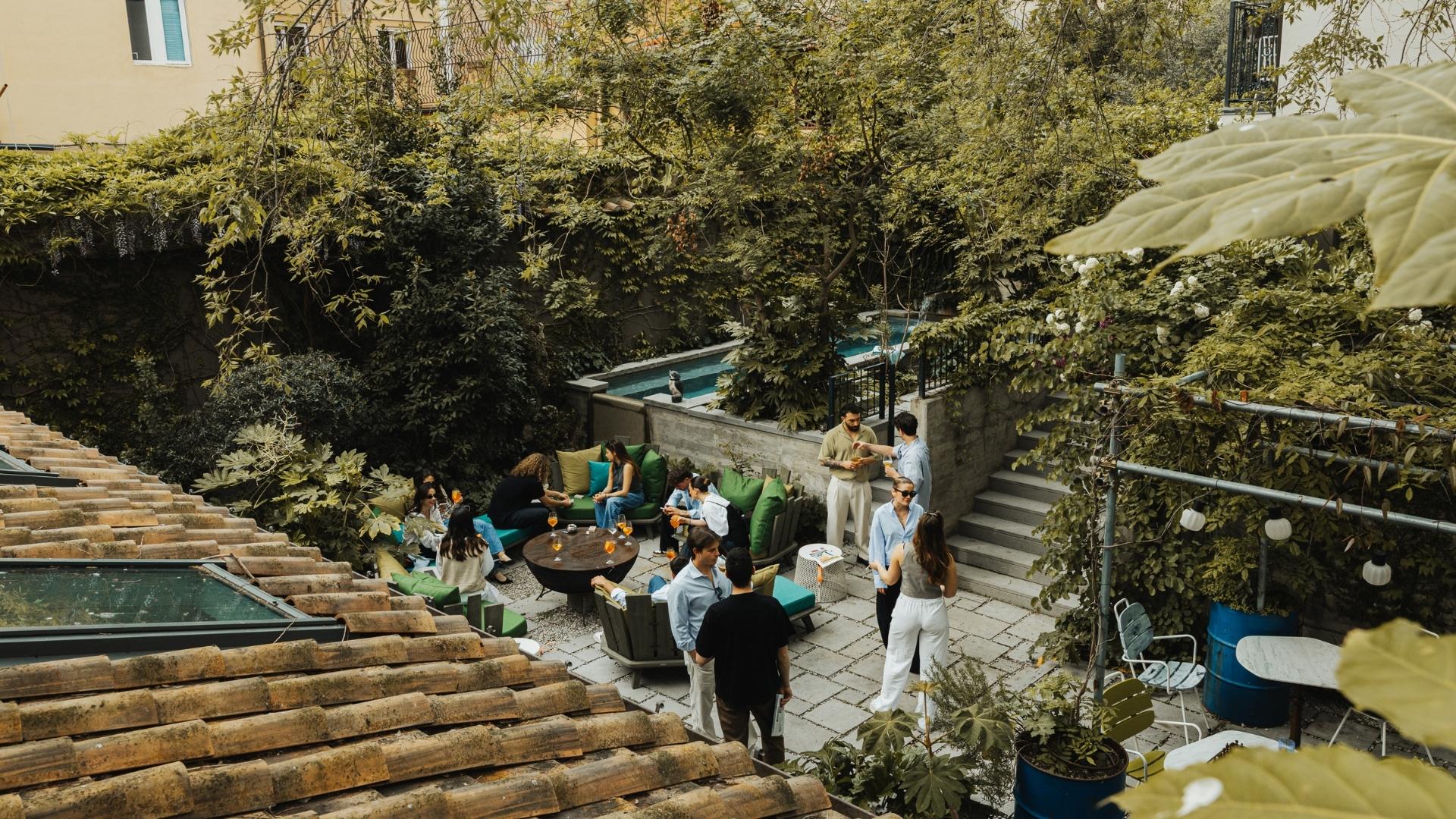 A lush garden gathering with people socializing around a round table. There are green cushions and a pool, creating a relaxed, inviting atmosphere.