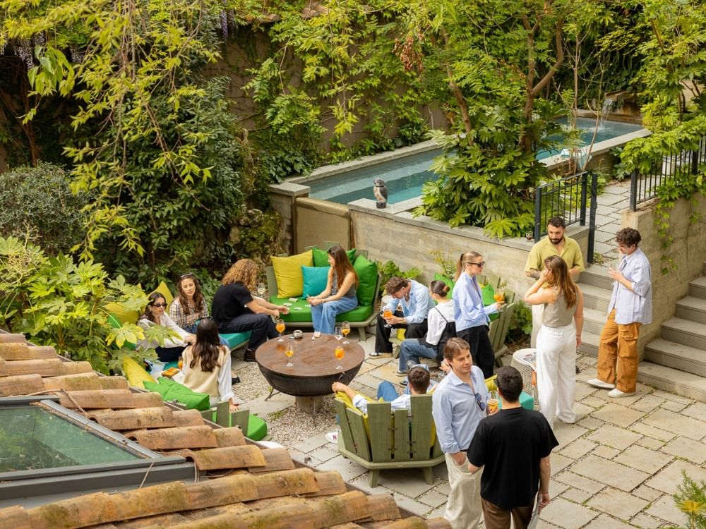 Group of people socializing outdoors in a lush garden patio with green cushions, round table, and a narrow water feature wall.