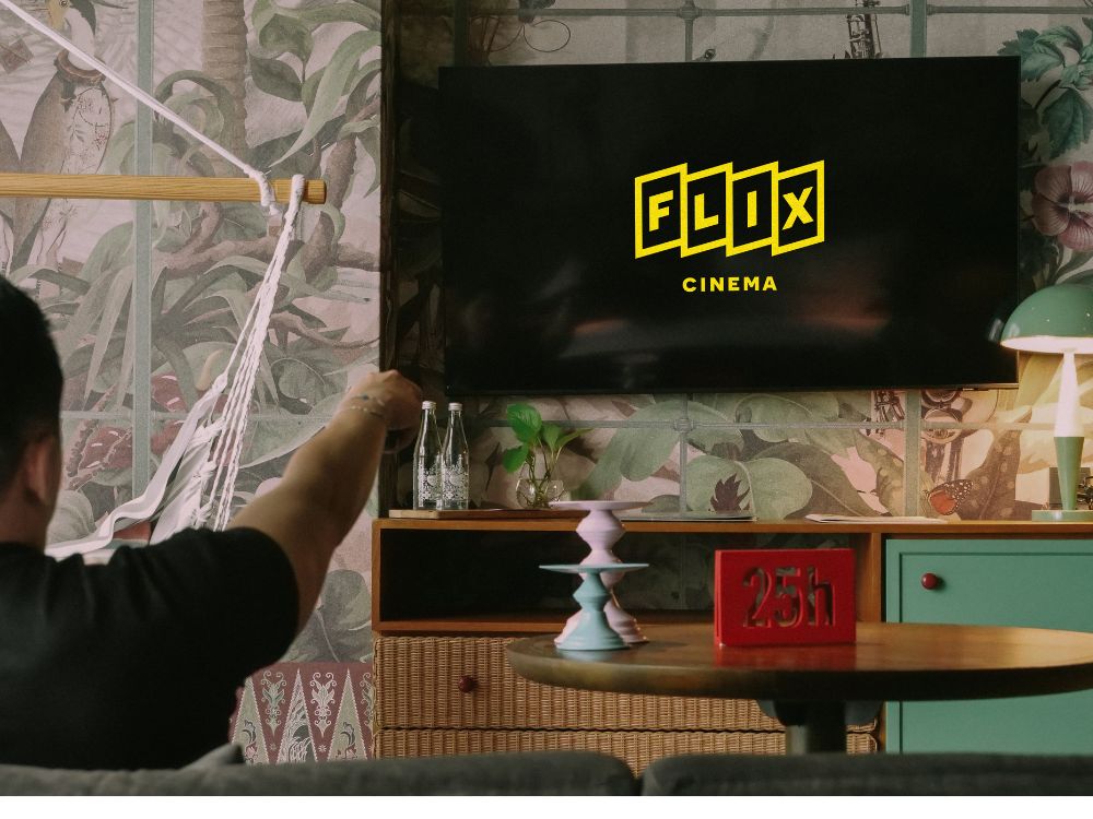 Cozy living room with a TV displaying "FLIX Cinema" logo. A person points a remote. Decorative wallpaper, hammock, and a table with a "25h" sign create a relaxed vibe.