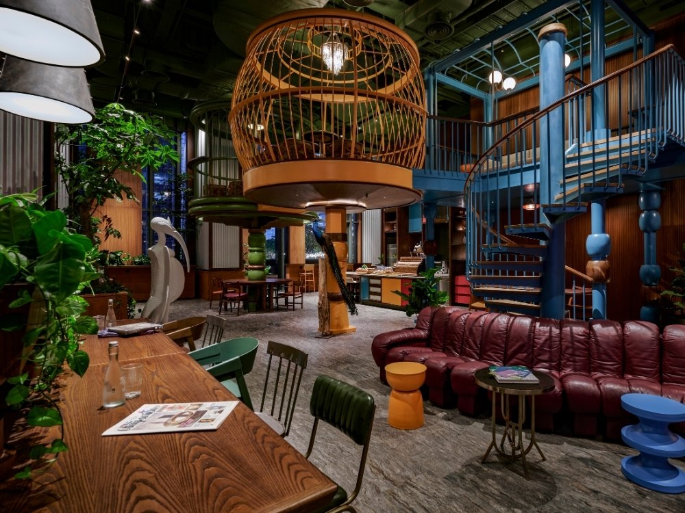 Eclectic indoor lounge with a large birdcage light fixture, spiral staircase, leather sofa, wooden table, and vibrant green plants.
