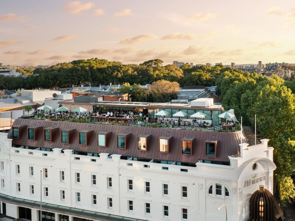 Aerial view of a rooftop bar on a historic white building with a brown roof, surrounded by trees, at sunset. People enjoy drinks under umbrellas.