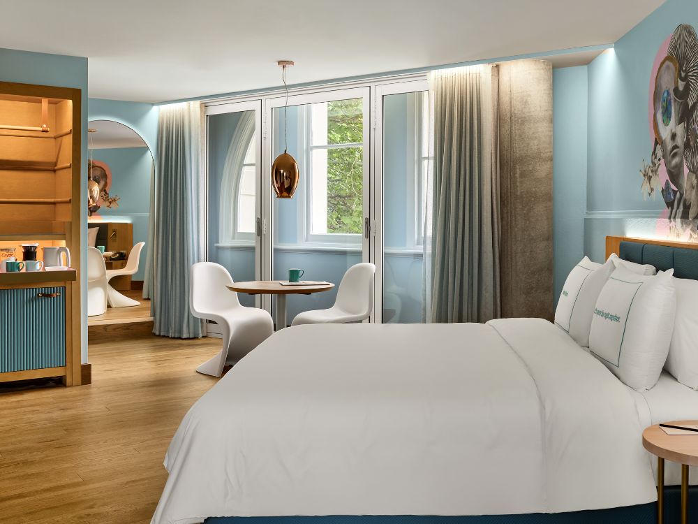 Bright hotel room with king bed, blue walls, round table with two white chairs, wooden floor, and large windows with curtains.