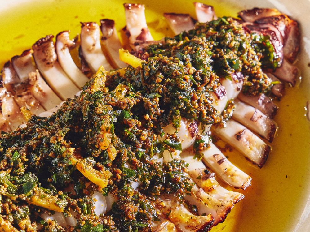 Grilled squid sliced and arranged in a fanned-out pattern, topped with a vibrant green and brown chimichurri sauce, all drizzled with golden oil.